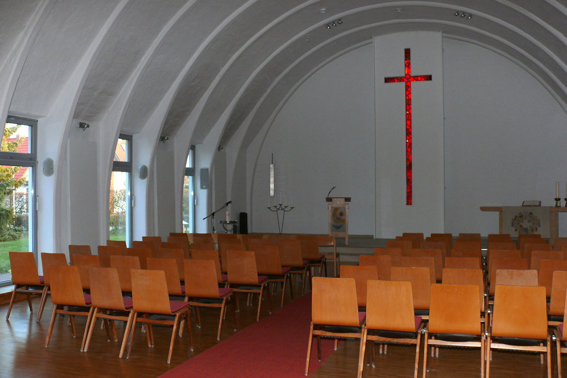 Stuhlreihen auf rotem Teppich / an der vorderen weißen Wand ist ein rotes Kreuz aus Glas zu sehen