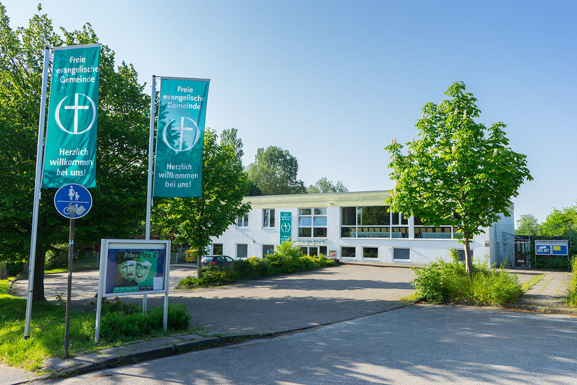 Ein weißes Flachdachgebäude im Hintergrund, davor freie Parkplätze / Im Vordergrund ein Schaukasten mit einem Plakat mit Masken und Schriftzug "Poker Face", links und rechts daneben grüne Fahnen mit weißem Schriftzug: Freie evangelische Gemeinde - Herzlich willkommen bei uns!