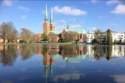 Im Vordergrund ist der Mühlenteich zu sehen, in welchen sich der der Dom spiegelt. Der Dom ist im Hintergrund zu sehen. Die Türme ragen in den nur leicht mit wolken bedeckten Himmel - Copyright: Ev.-Luth. Kirchenkreis Lübeck-Lauenburg