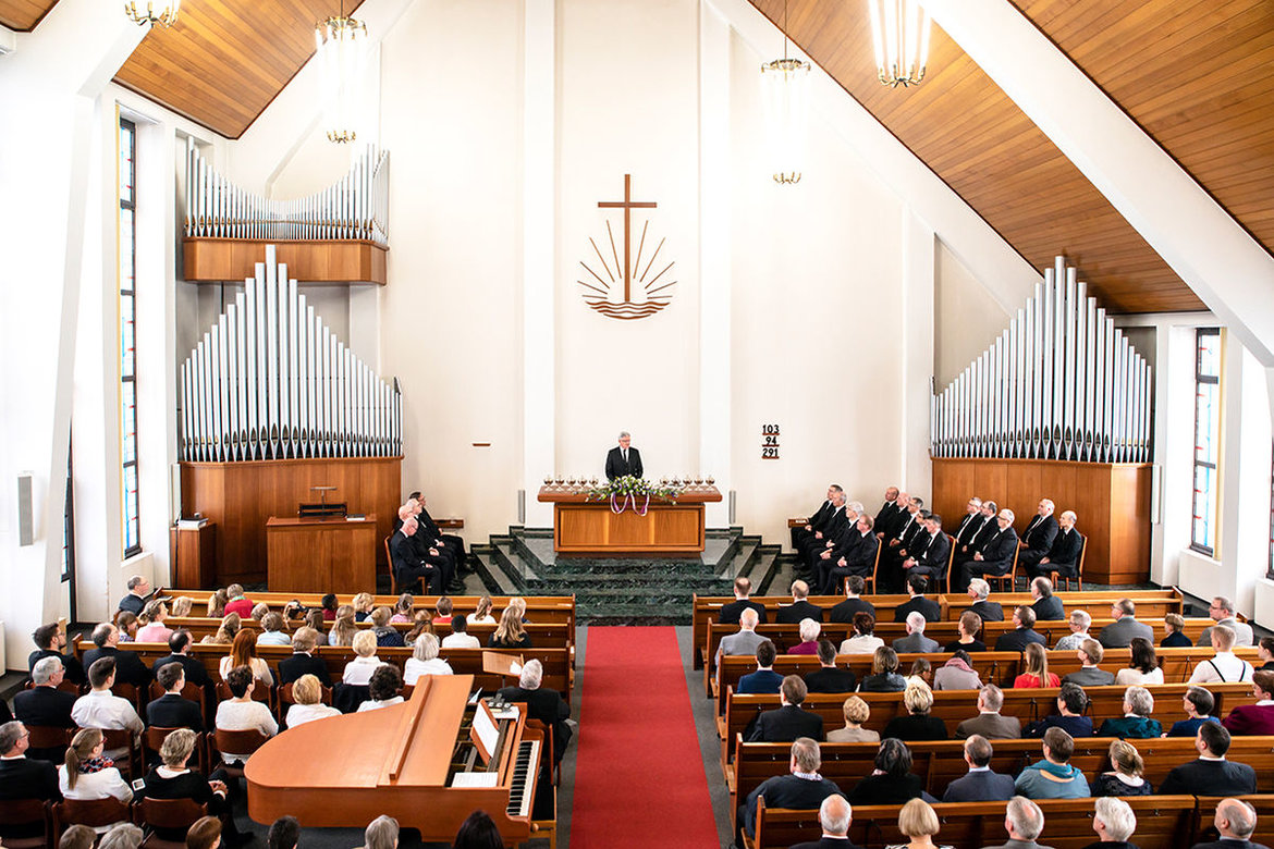 Im Vordergrund holzbänke mit Gottesdienstbesuchern / Im Hintergrund links und rechts hellbraune Orgeln mit Orgelpfeifen / In der Mitte der Altar wo der Pastor hinter steht, links und rechts daneben Stuhlreihen mit sitzenden Männern.